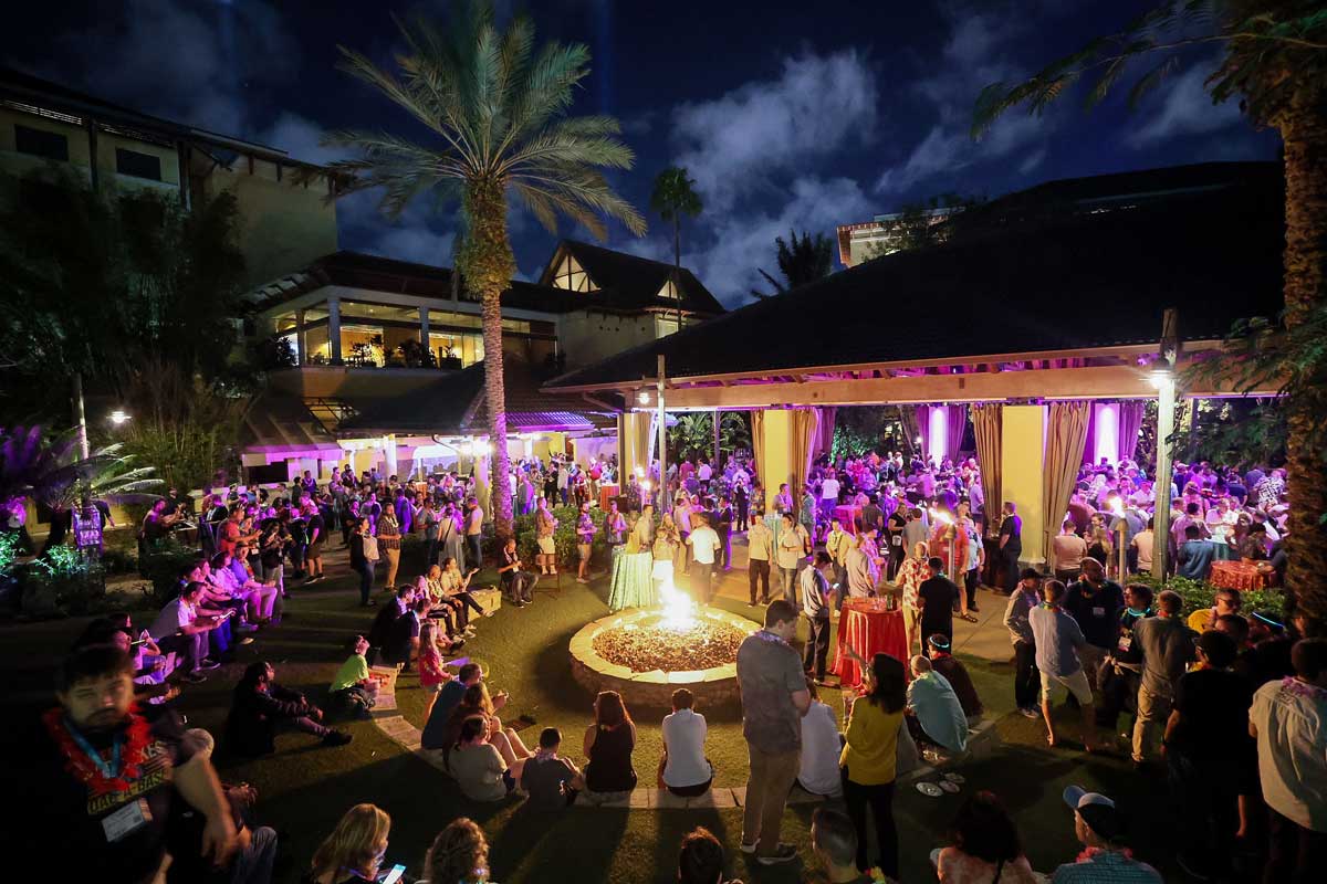 Live! 360 attendees gathered outdoors at night around a fire pit under palm trees, lit in purple, at the Dessert Luau.